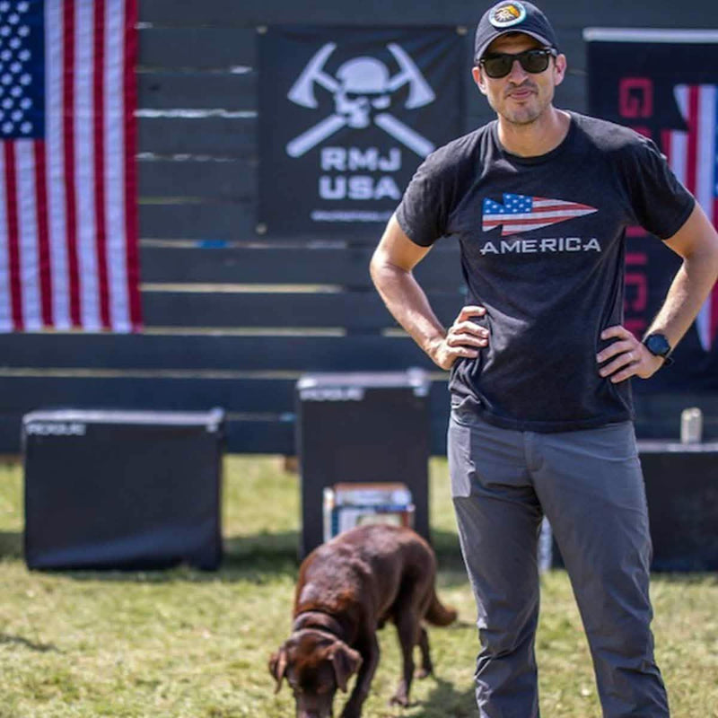 Man in GORUCK America shirt outdoors with American flag and dog at rucking event