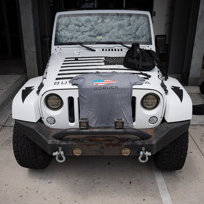The GORUCK Flag Spearhead Tee - Unisex Tri-BlendX in gray and a black bag with the GORUCK spearhead are displayed on the hood of a white Jeep with an American flag decal.