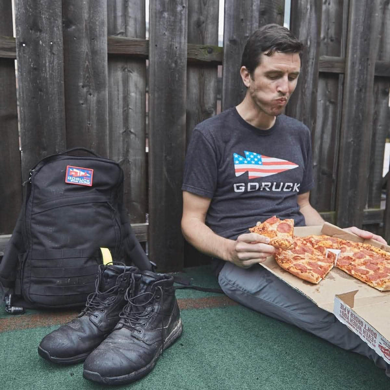 Man in GORUCK USA t-shirt eating pepperoni pizza outdoors, with backpack and boots nearby