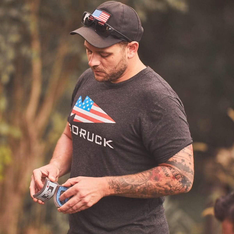 Man wearing GORUCK USA t-shirt and hat outdoors, holding rucking patches, rugged style