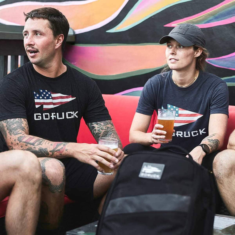 Two people wearing GORUCK American flag shirts, holding drinks, sitting with a GORUCK backpack.