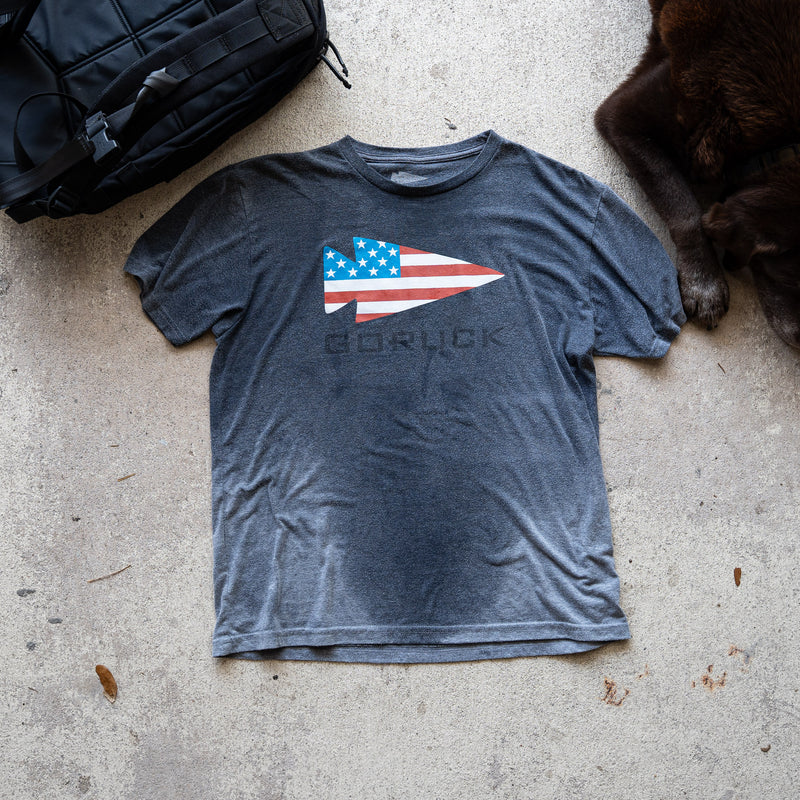 The GORUCK Flag Spearhead Tee - Unisex Tri-BlendX, a gray premium T-shirt featuring an American flag and Special Forces-inspired spearhead, is shown on concrete next to a black bag and a dog's paw.