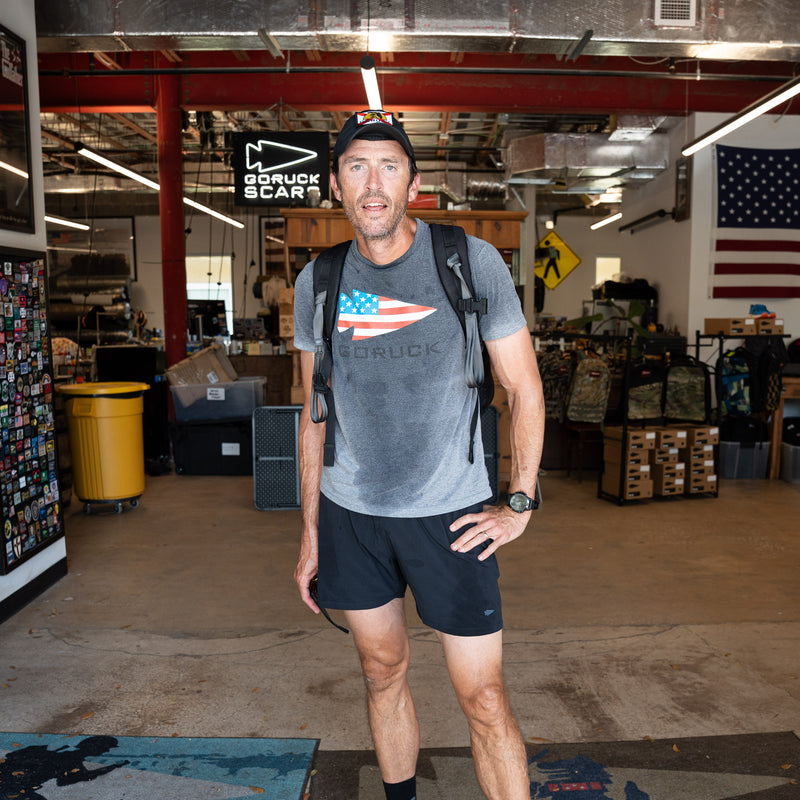 A man in the GORUCK Flag Spearhead Tee - Unisex Tri-BlendX, featuring the iconic spearhead and backpack, stands in a gear shop, looking tired but happy.