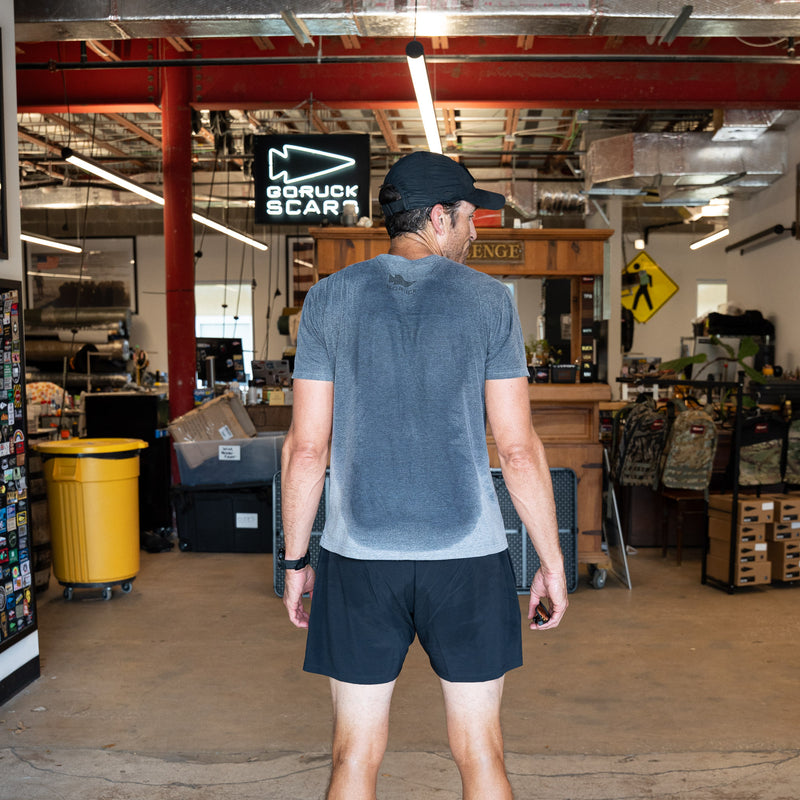 A man in athletic gear, wearing the GORUCK Flag Spearhead Tee - Unisex Tri-BlendX with a visible sweat stain and logo on his back, stands inside a gear store facing away from the camera.