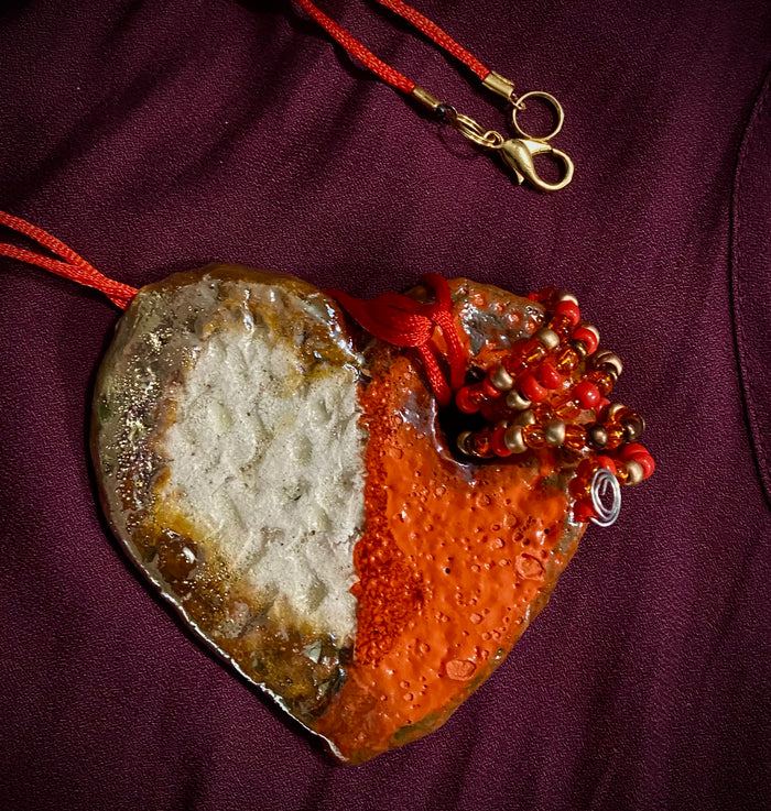 Have A Heart! Each heart pendant is handmade with love! It is 3"x 3"and weighs approx. 3ozs. This pendant has a red,orange, white and copper metallic raku glazes that renders a unique translucent  patina. The heart has a textured pattern . Both sides are  are different and equally beautiful! There is a spiral of red mini beads on copper in the center. This pendant has a nice 12" red adjustable rattail cord!