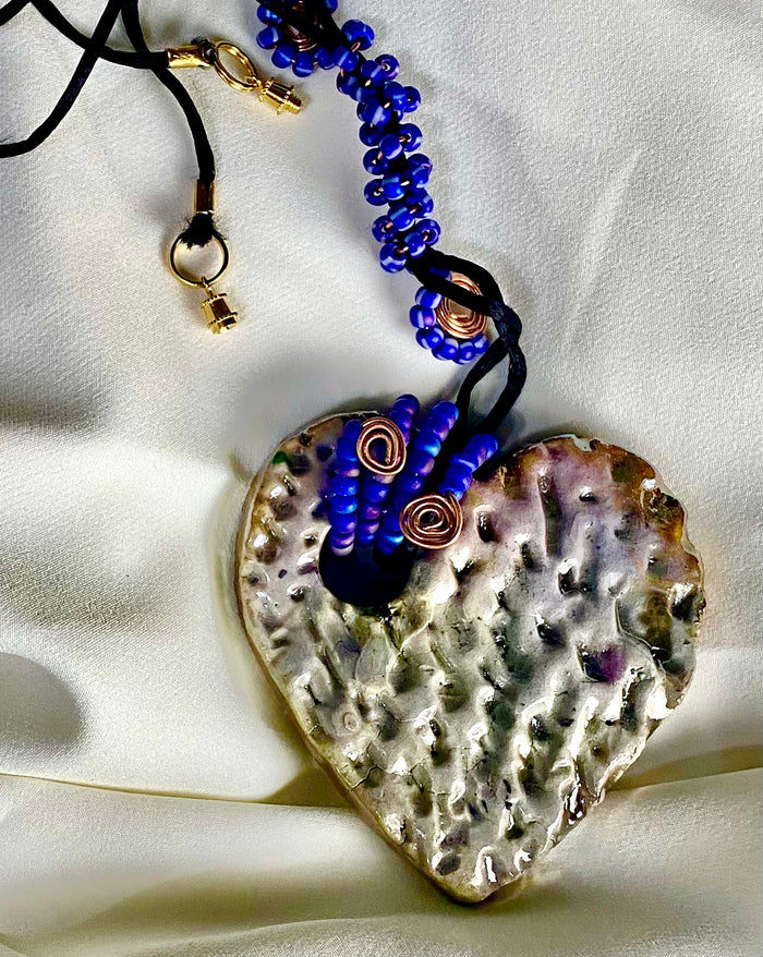 Have A Heart ! Each heart pendant is handmade with love! It is 3"x 3" and weighs approx. 3ozs. This pendant has a white metallic raku glazes that renders a unique translucent  patina. The heart has a textured pattern . It holds a spiral of blue mini beads on a spiral copper wire. This pendant has a nice 12" black suede cord!