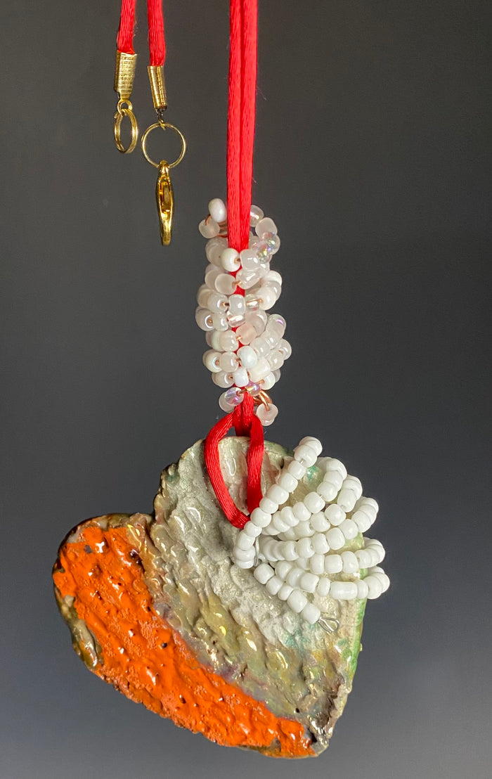 Have A Heart ! Each heart pendant is handmade with love! It is 3"x 3" and weighs approx. 3ozs. This pendant has a orange gold metallic raku glazes that renders a unique translucent  patina. The heart has a textured pattern . It holds a spiral of white and clear mini beads on a spiral copper wire. This pendant has a nice 12" red suede cord!