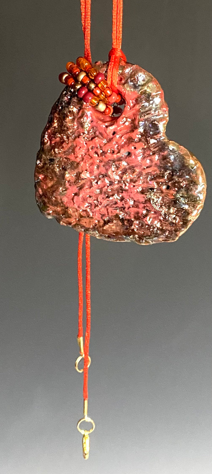 Have A Heart! Each heart pendant is handmade with love! It is 3"x 3" and weighs approx. 3ozs. This pendant has a red violet and gold metallic raku glazes that renders a unique translucent  patina. The heart has a textured pattern . Both sides are  are different and equally beautiful! It holds a spiral of red mini beads on a spiral copper wire. This pendant has a nice 12" red rattail cord!
