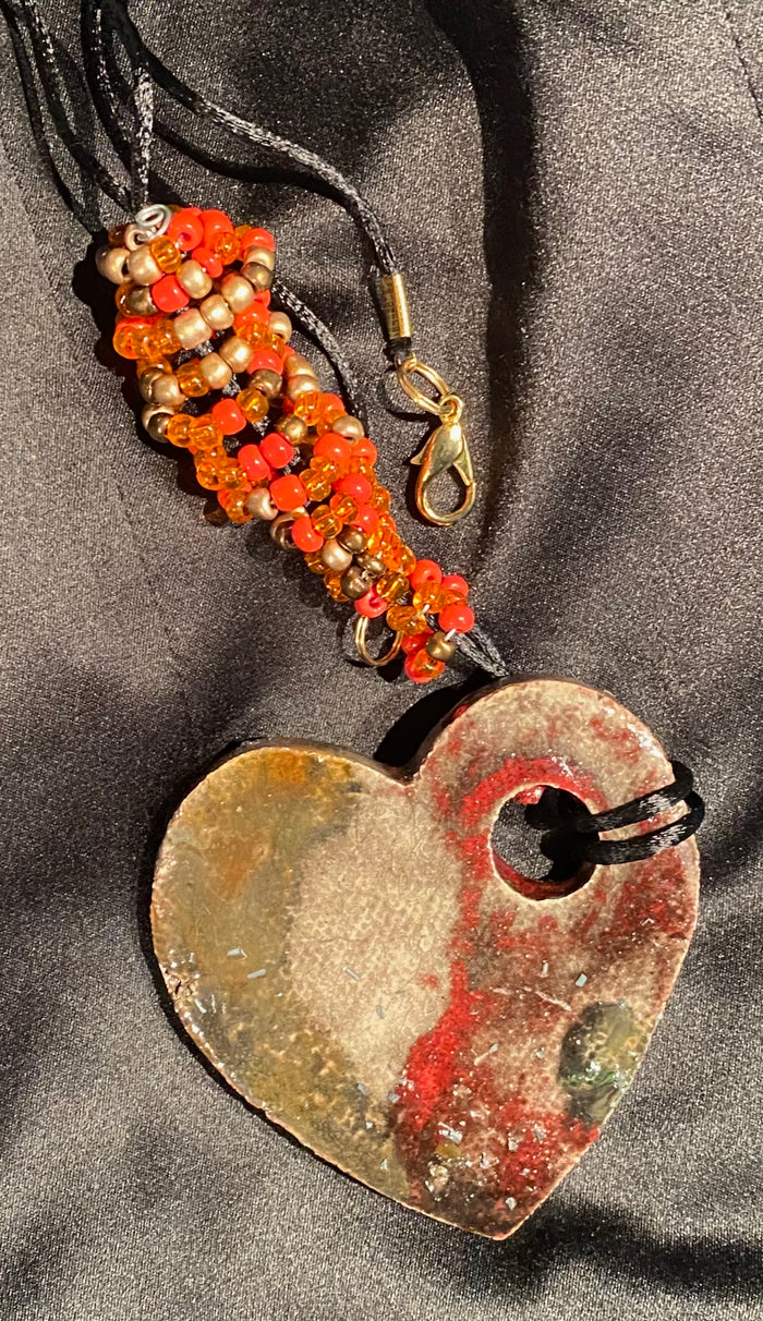 Have A Heart! Each heart pendant is handmade with love! It is 3"x 3"and weighs approx. 3ozs. This pendant has a red violet and gold metallic raku glazes that renders a unique translucent  patina. The heart has a textured pattern . Both sides are  are different and equally beautiful! It holds a spiral of red ad amber mini beads on a spiral copper wire. This pendant has a nice 12" black rattail cord!
